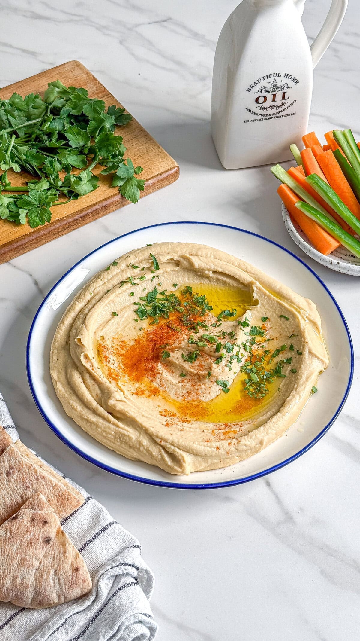 A plate of easy creamy hummus garnished with olive oil, paprika, and herbs sits on a marble surface, surrounded by pita bread, fresh vegetables, herbs on a cutting board, and a ceramic oil bottle.