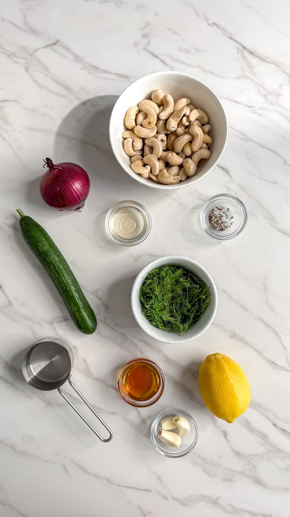 Overhead view of ingredients for a creamy vegan cucumber salad on a white marble surface: cashews, red onion, cucumber, dill, lemon, garlic cloves, a measuring cup, and small bowls of vinegar, oil, and pepper.