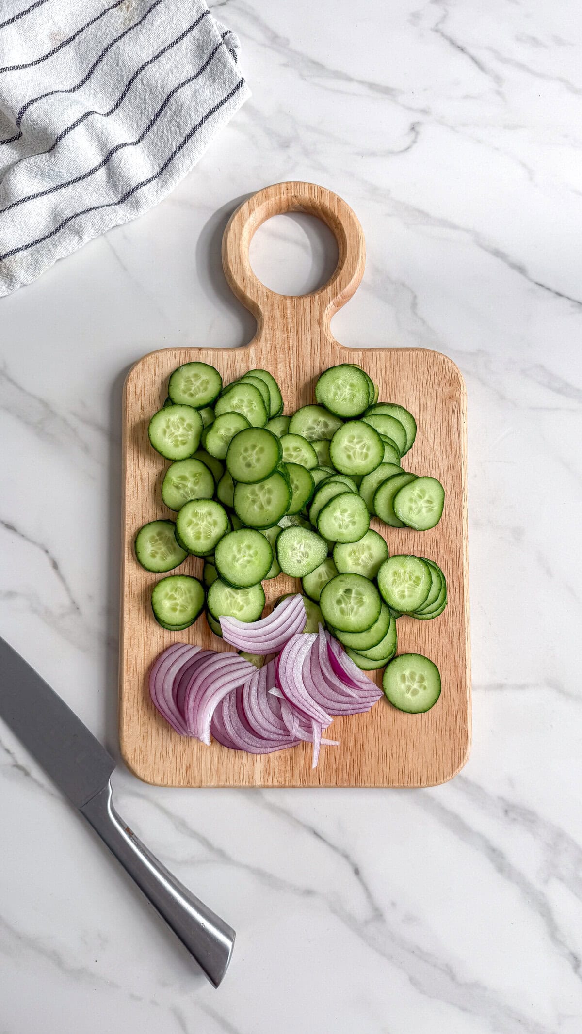 A wooden cutting board with sliced cucumbers and red onions sits on a marble countertop, ready for a creamy vegan cucumber salad. A knife lies next to the board and a striped kitchen towel is in the corner.