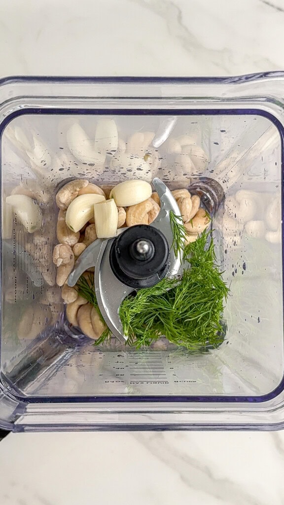 A top-down view of a blender containing raw cashews, garlic cloves, and fresh dill on a white marble countertop—perfect ingredients for a creamy vegan cucumber salad dressing.