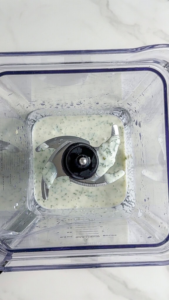 Top-down view of a blender containing a small amount of creamy, speckled sauce for a vegan cucumber salad, with metal blades visible and some mixture splattered on the sides.