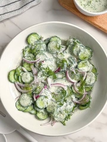 A white bowl filled with creamy vegan cucumber salad, featuring sliced cucumbers, red onion, and fresh dill coated in a luscious herb dressing. A small bowl of extra dressing and a striped kitchen towel rest on the marble surface nearby.