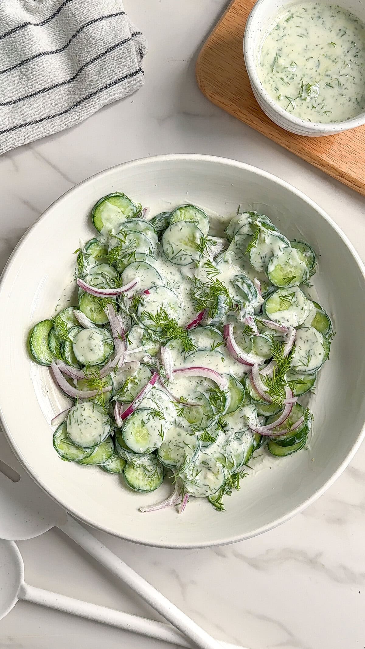 A white bowl filled with creamy vegan cucumber salad, featuring sliced cucumbers, red onion, and fresh dill coated in a luscious herb dressing. A small bowl of extra dressing and a striped kitchen towel rest on the marble surface nearby.