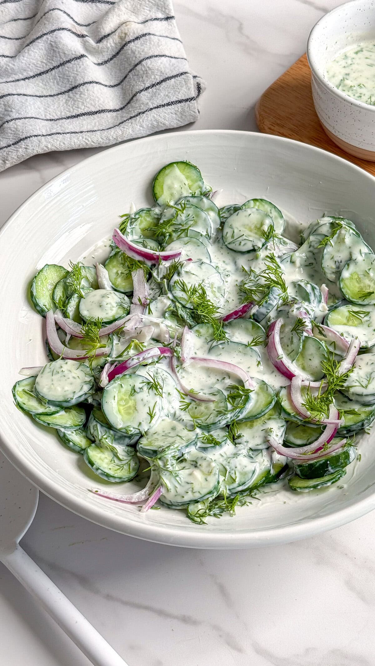 A white bowl filled with creamy vegan cucumber salad, featuring sliced cucumbers, red onions, fresh dill, and a luscious dressing. A striped towel and a small bowl of extra dressing are in the background.