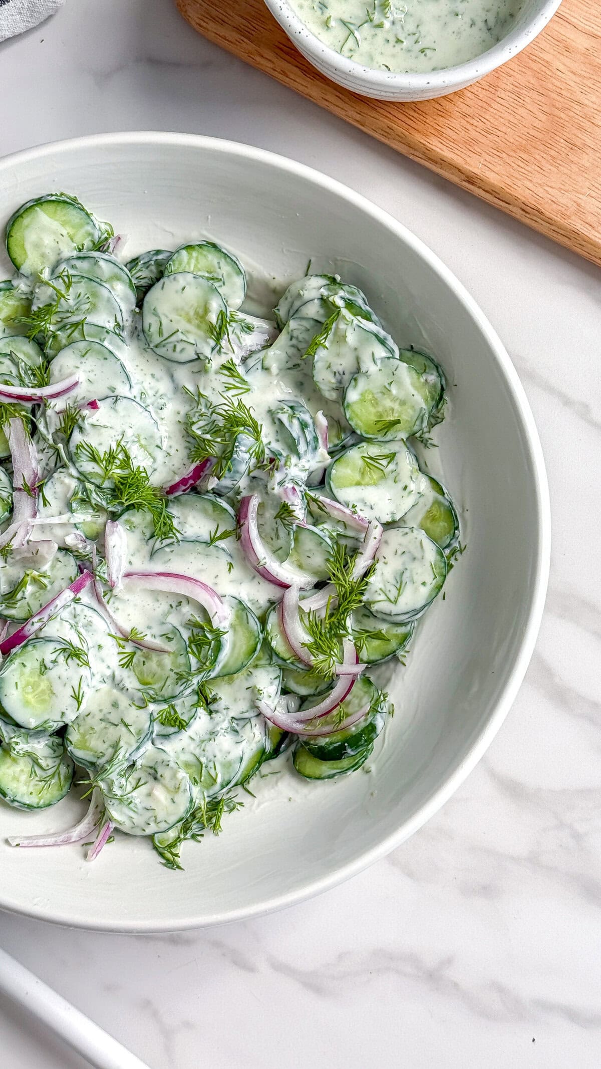 A white bowl filled with creamy vegan cucumber salad mixed with sliced red onions, fresh dill, and creamy dressing sits on a marble surface. A small bowl of extra dressing is partially visible in the background.