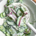 A close-up of a white spoon holding creamy vegan cucumber salad with sliced cucumbers, red onions, and fresh dill above a bowl of the same salad. A small bowl of dressing is visible in the background.