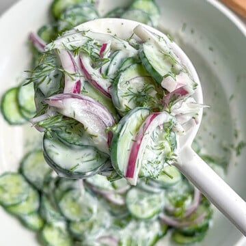 A close-up of a white spoon holding creamy vegan cucumber salad with sliced cucumbers, red onions, and fresh dill above a bowl of the same salad. A small bowl of dressing is visible in the background.