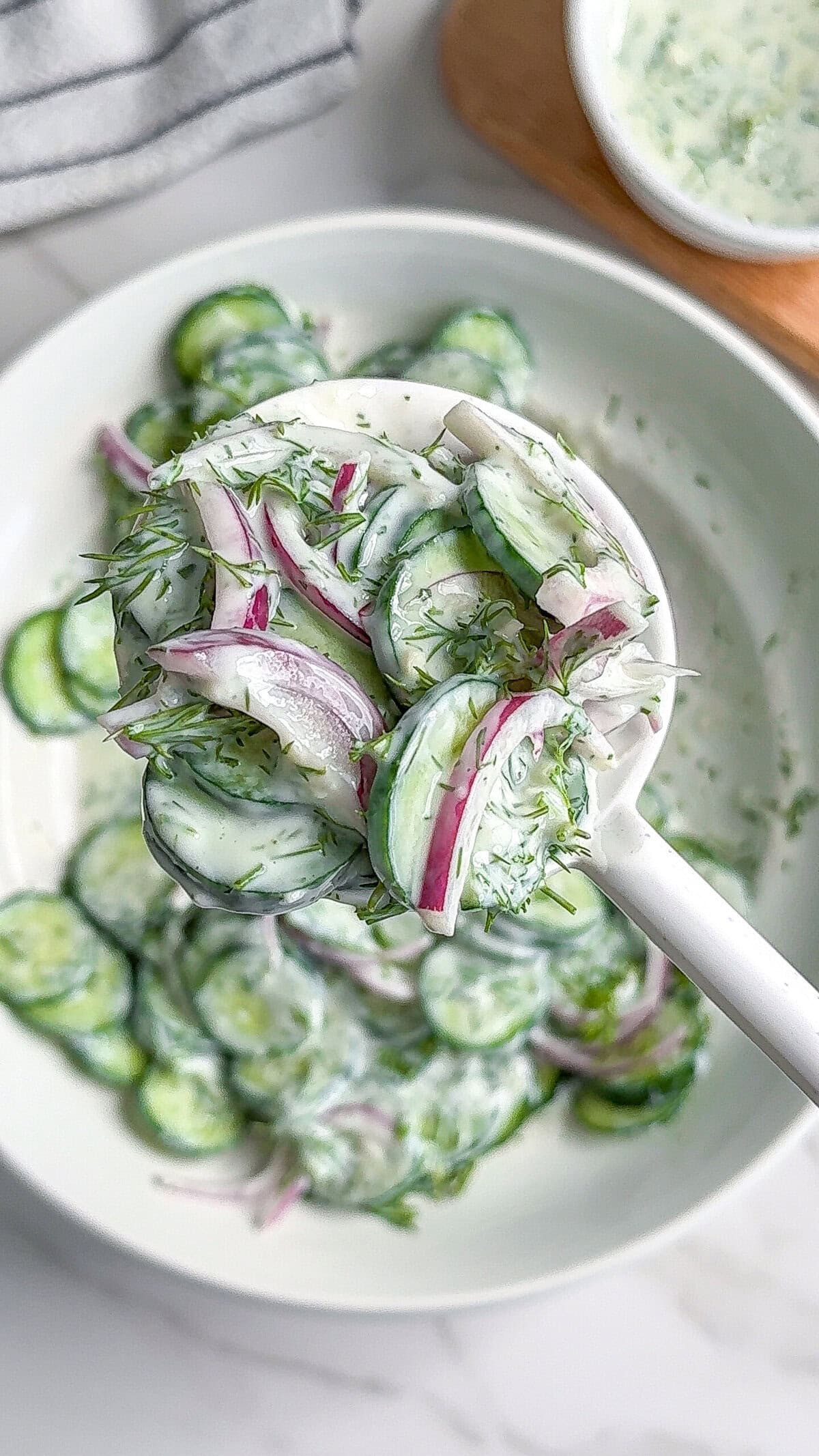 A close-up of a white spoon holding creamy vegan cucumber salad with sliced cucumbers, red onions, and fresh dill above a bowl of the same salad. A small bowl of dressing is visible in the background.
