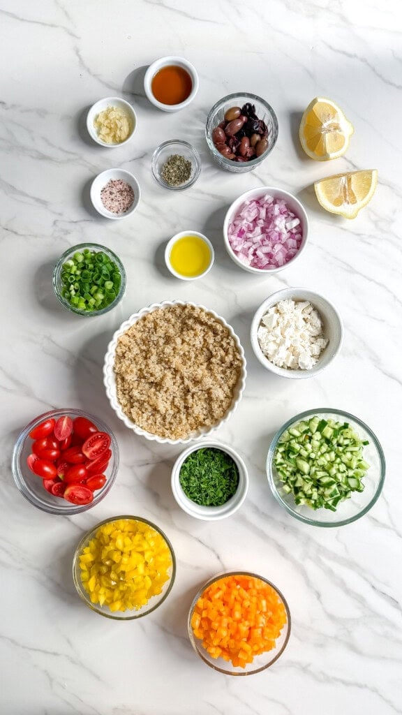 Overhead view of small bowls filled with ingredients for a vegan Mediterranean quinoa salad, including chopped red onion, green onion, cherry tomatoes, cucumbers, bell peppers, olives, feta cheese, parsley, lemon wedges, oil, vinegar, salt, and pepper.