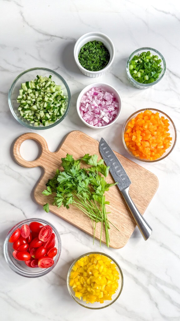 Chopped veggies in glass bowls for a vegan Mediterranean quinoa salad, parsley on a wooden cutting board with a knife—all arranged on a white marble surface. Tomatoes, cucumbers, onions, bell peppers, green onions, and parsley are visible.