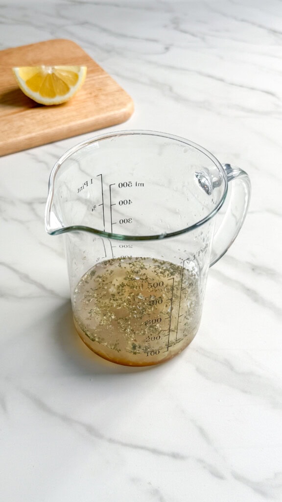 A glass measuring cup with a small amount of liquid dressing mixed with herbs sits on a marble surface—perfect for drizzling over your vegan Mediterranean quinoa salad. In the background, a wooden board holds a lemon wedge.