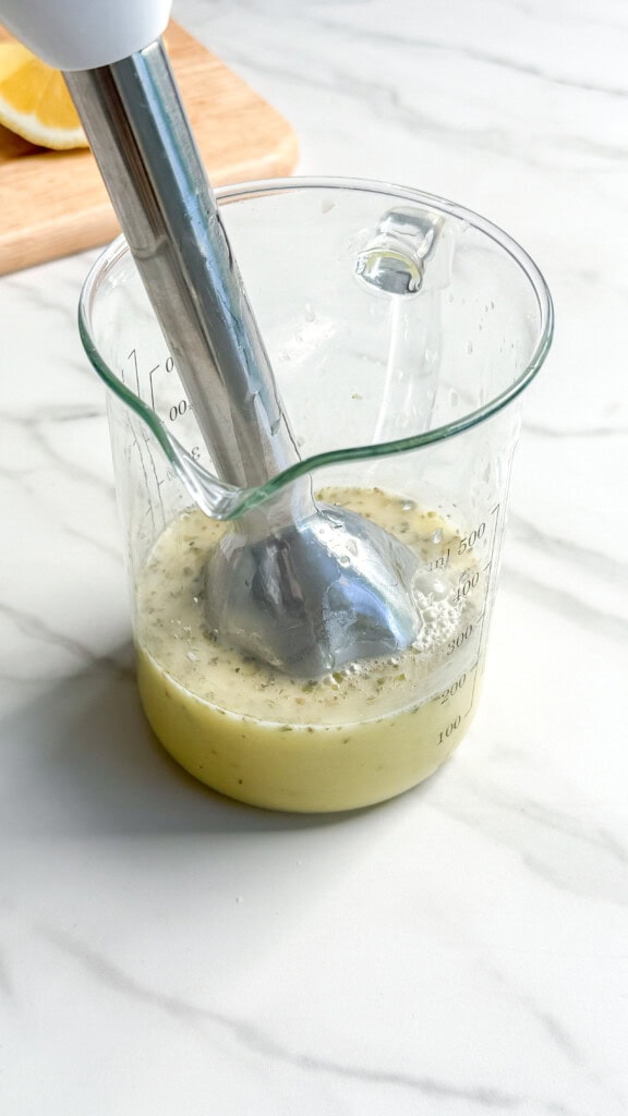 An immersion blender blending a light green mixture with herbs in a glass measuring cup on a white marble countertop, perfect for dressing a vegan Mediterranean quinoa salad, with a wooden board and a lemon in the background.