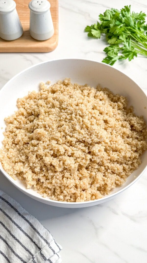 A white bowl filled with cooked quinoa sits on a white countertop, ready for a vibrant vegan Mediterranean quinoa salad. Fresh parsley, a striped towel, and salt and pepper shakers add color and style to the scene.