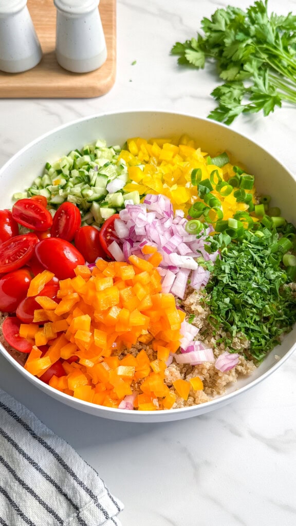 A bowl filled with chopped yellow bell pepper, cherry tomatoes, cucumber, red onion, green onion, parsley, and quinoa creates a vibrant vegan Mediterranean quinoa salad. Fresh parsley and salt shakers are nearby on the white countertop.
