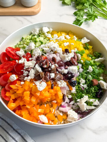 A large white bowl filled with colorful chopped vegetables, including yellow and orange bell peppers, cherry tomatoes, cucumber, red onion, olives, and crumbled feta cheese, creates a vibrant vegan Mediterranean quinoa salad on a marble countertop.