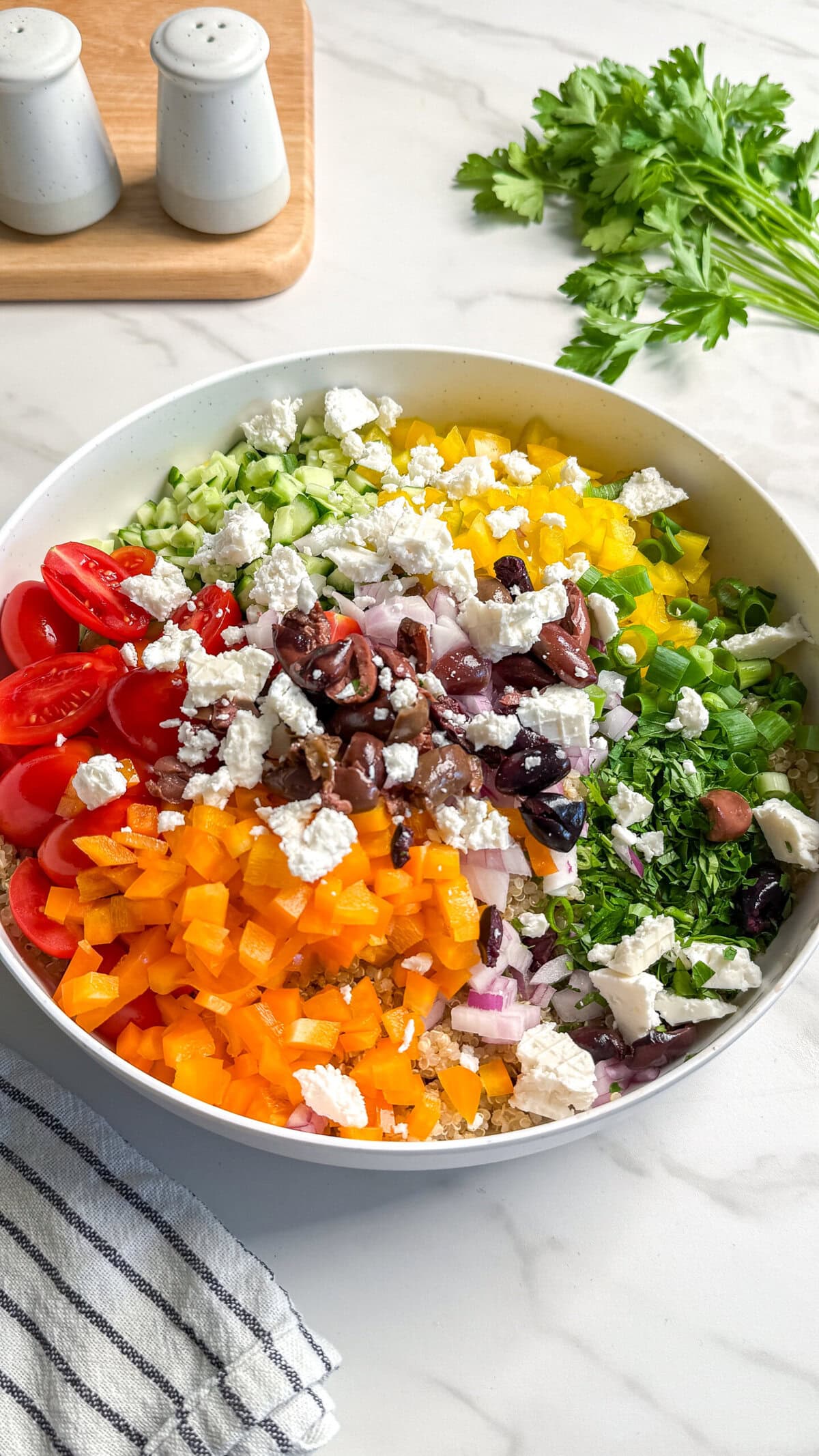 A large white bowl filled with colorful chopped vegetables, including yellow and orange bell peppers, cherry tomatoes, cucumber, red onion, olives, and crumbled feta cheese, creates a vibrant vegan Mediterranean quinoa salad on a marble countertop.