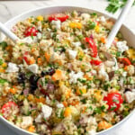A bowl of colorful vegan Mediterranean quinoa salad with chopped vegetables, cherry tomatoes, cucumbers, fresh herbs, and two serving spoons. Salt and pepper shakers and parsley are in the background.
