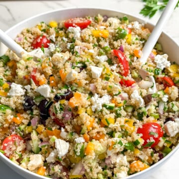 A bowl of colorful vegan Mediterranean quinoa salad with chopped vegetables, cherry tomatoes, cucumbers, fresh herbs, and two serving spoons. Salt and pepper shakers and parsley are in the background.