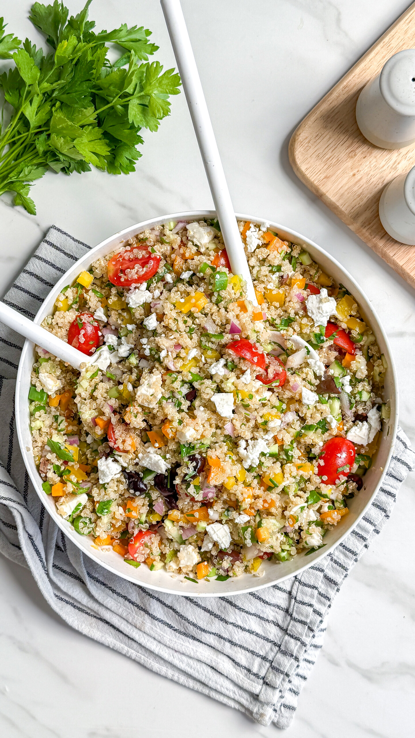 A bowl of vegan Mediterranean quinoa salad with cherry tomatoes, yellow peppers, cucumbers, olives, red onion, and feta cheese sits with two serving spoons. Fresh parsley and salt and pepper shakers are nearby on a marble countertop.
