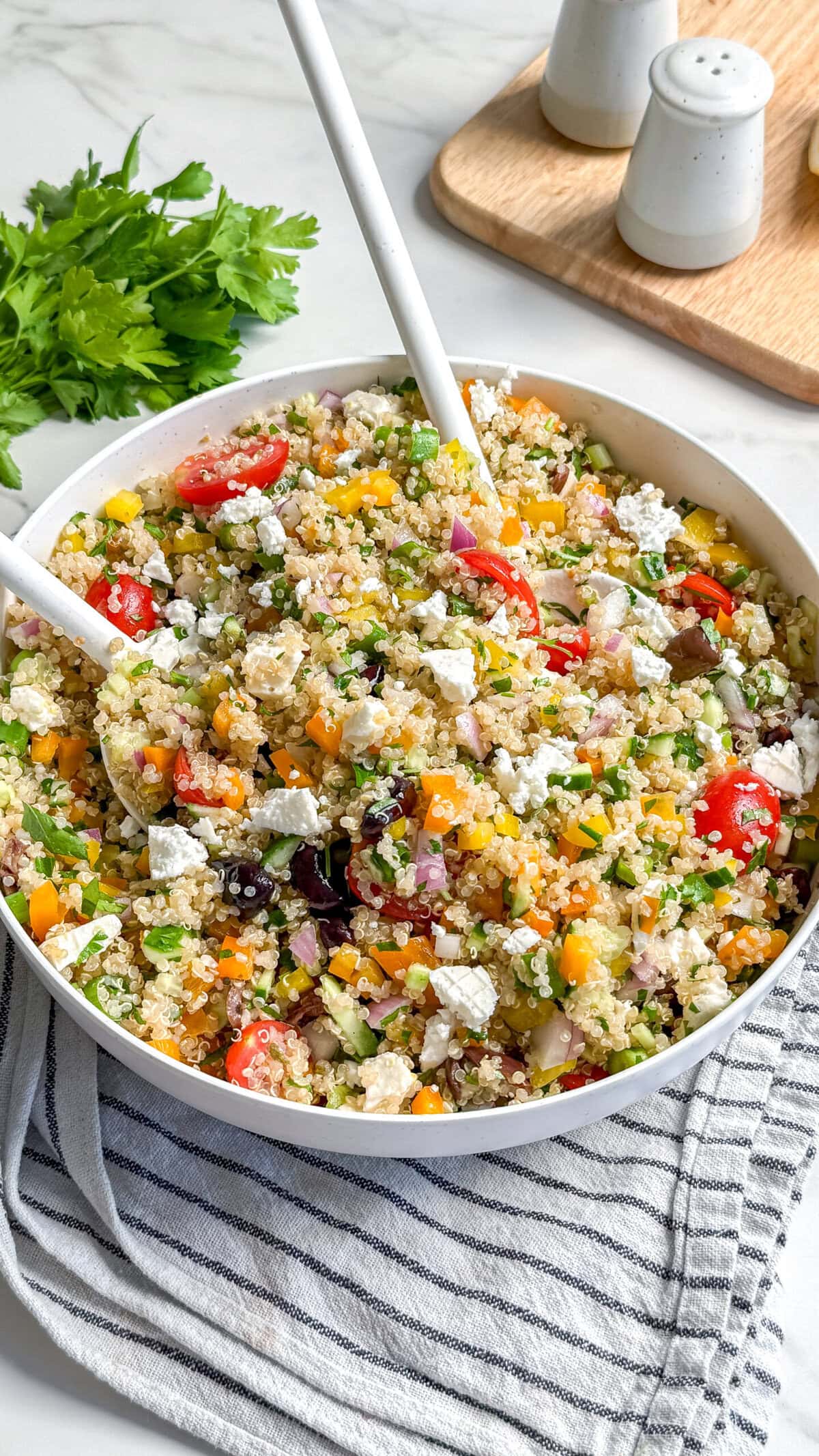 A large bowl of vibrant vegan Mediterranean quinoa salad with chopped vegetables, cherry tomatoes, herbs, and two serving spoons. Fresh parsley, salt, and pepper shakers are in the background.