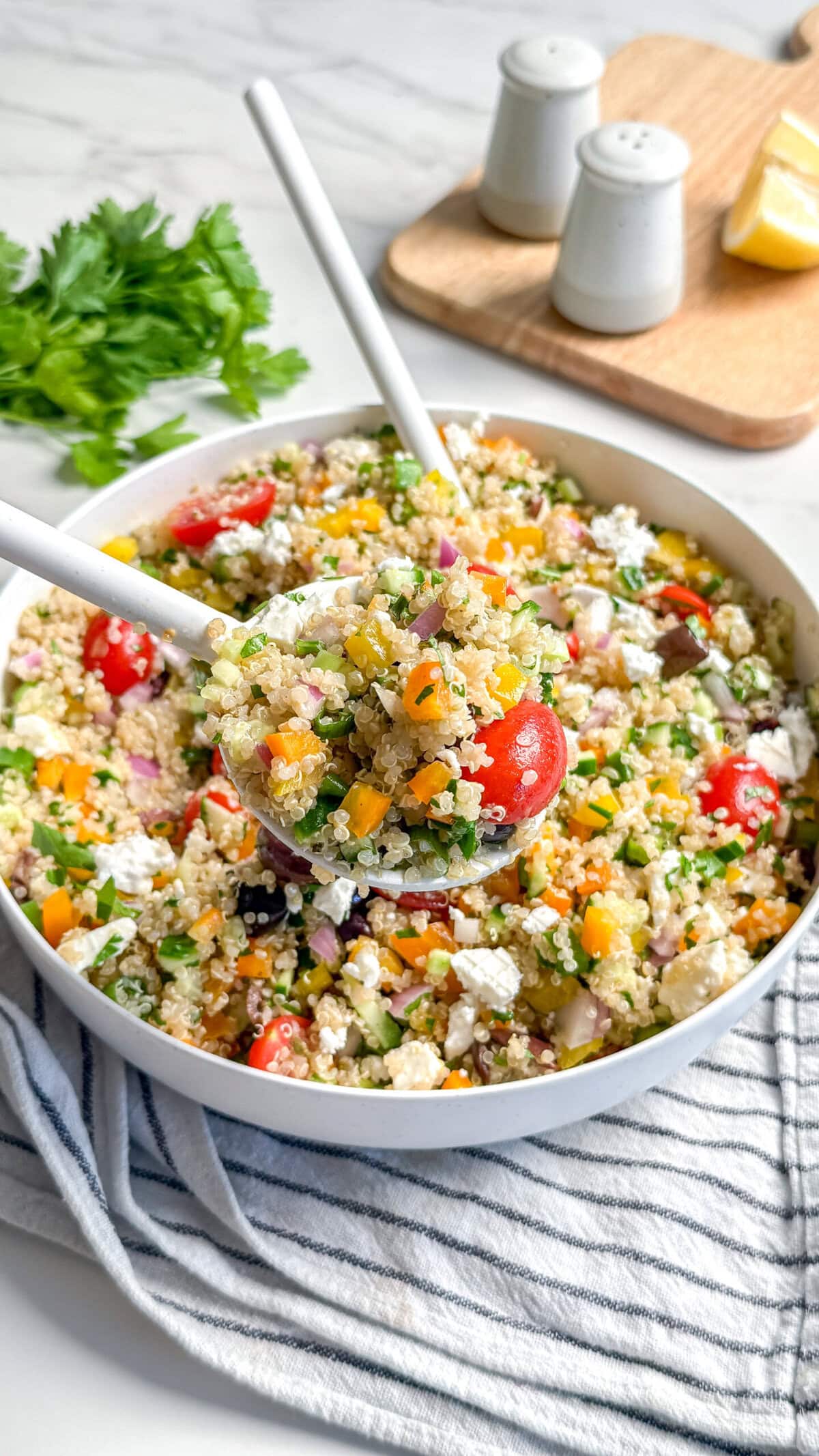 A bowl of vegan Mediterranean quinoa salad with chopped vegetables, cherry tomatoes, red onion, yellow bell pepper, herbs, and crumbled feta cheese. Lemon wedges and salt and pepper shakers sit in the background beside serving spoons.