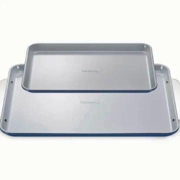 Two rectangular, light gray nonstick baking sheets—one a classic half-sheet stacked on top of a larger baking sheet—both with curved edges and visible handles, set on a plain light background.