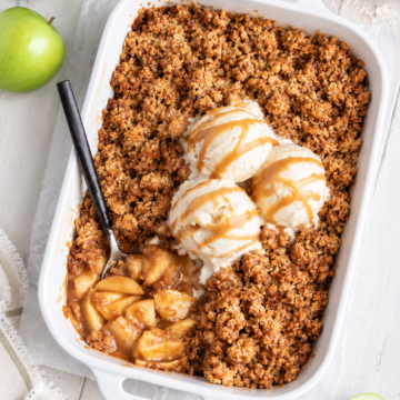 A white baking dish with Vegan Apple Crisp, topped with three scoops of vanilla ice cream and caramel sauce. A spoon rests in the dish, revealing baked apples. Green apples are on the table nearby.