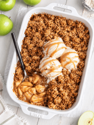 A white baking dish with Vegan Apple Crisp, topped with three scoops of vanilla ice cream and caramel sauce. A spoon rests in the dish, revealing baked apples. Green apples are on the table nearby.