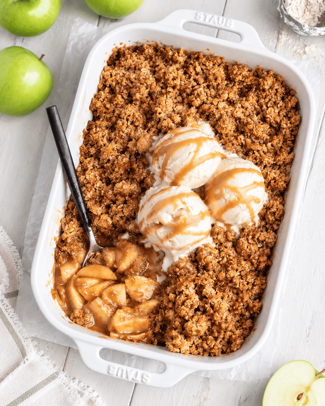 A white baking dish with Vegan Apple Crisp, topped with three scoops of vanilla ice cream and caramel sauce. A spoon rests in the dish, revealing baked apples. Green apples are on the table nearby.
