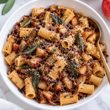A white bowl of rigatoni pasta with browned bits, apple pieces, fresh sage leaves, and grated cheese on top. A gold spoon rests in the bowl. Nearby on the marble surface are apples, sage leaves, a small bowl of cheese—and all the comforting flavors reminiscent of a Vegan Apple Crisp.
