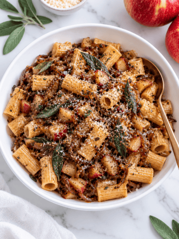 A white bowl of rigatoni pasta with browned bits, apple pieces, fresh sage leaves, and grated cheese on top. A gold spoon rests in the bowl. Nearby on the marble surface are apples, sage leaves, a small bowl of cheese&mdash;and all the comforting flavors reminiscent of a Vegan Apple Crisp.