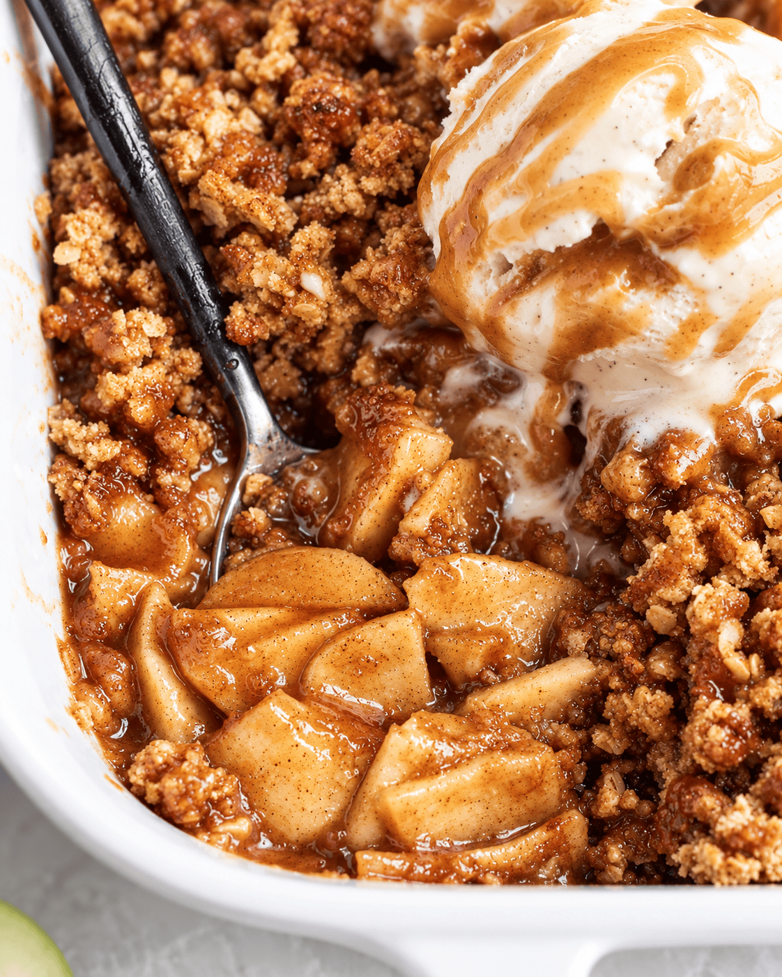 Close-up of Vegan Apple Crisp in a white dish, showing baked cinnamon apples, a crumbly brown topping, and a scoop of melting vanilla ice cream with a drizzle of caramel sauce. A spoon rests in the dessert.