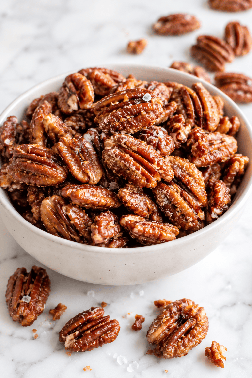 A white bowl filled with healthy candied pecans sits on a marble surface, with a few sugared pecans and grains of sea salt scattered around the bowl.