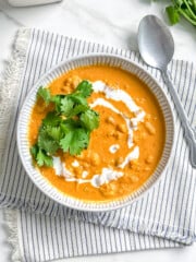 A bowl of vegan pumpkin curry topped with a swirl of cream and fresh cilantro, served with a spoon on a striped napkin.