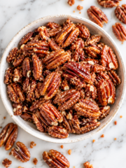 A bowl filled with healthy candied pecans sits on a marble surface, with a few pecans and crumbs scattered around it.