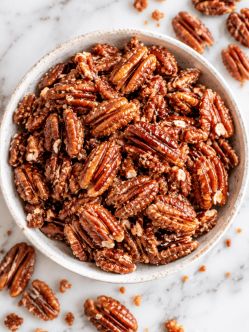 A bowl filled with healthy candied pecans sits on a marble surface, with a few pecans and crumbs scattered around it.