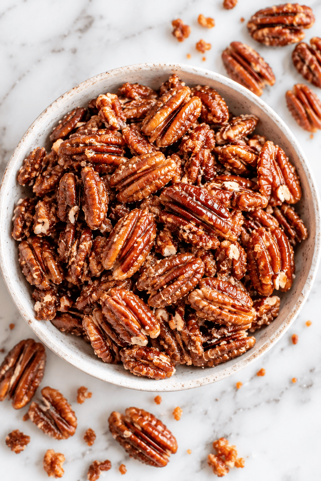 A bowl filled with healthy candied pecans sits on a marble surface, with a few pecans and crumbs scattered around it.