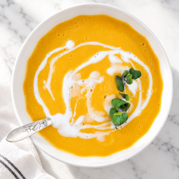 A bowl of creamy orange soup, this Easy Vegan Pumpkin Soup features a swirl of white cream and a garnish of green microgreens, served with a spoon on a white marble surface.