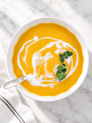 A bowl of creamy orange soup, this Easy Vegan Pumpkin Soup features a swirl of white cream and a garnish of green microgreens, served with a spoon on a white marble surface.
