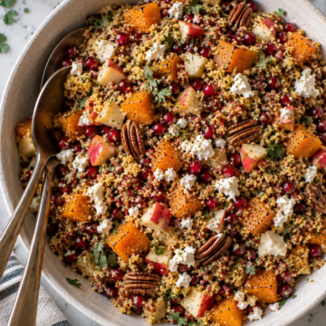 A bowl of Butternut Squash and Apple Quinoa Salad with roasted butternut squash, apples, pecans, pomegranate seeds, feta cheese, and fresh herbs. Two serving spoons rest inside the bowl, and a striped napkin is nearby.