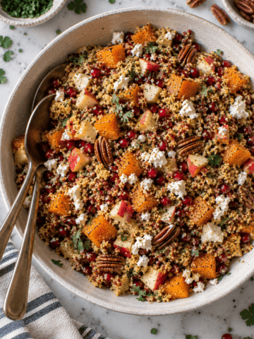 A bowl of Butternut Squash and Apple Quinoa Salad with roasted butternut squash, apples, pecans, pomegranate seeds, feta cheese, and fresh herbs. Two serving spoons rest inside the bowl, and a striped napkin is nearby.