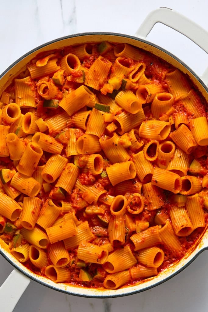 A white pot filled with rigatoni pasta mixed in a bright red tomato sauce with visible pieces of zucchini, all set on a white marble surface—perfect for an easy vegan pasta bake if you’re searching for how to cook pasta bake in oven.