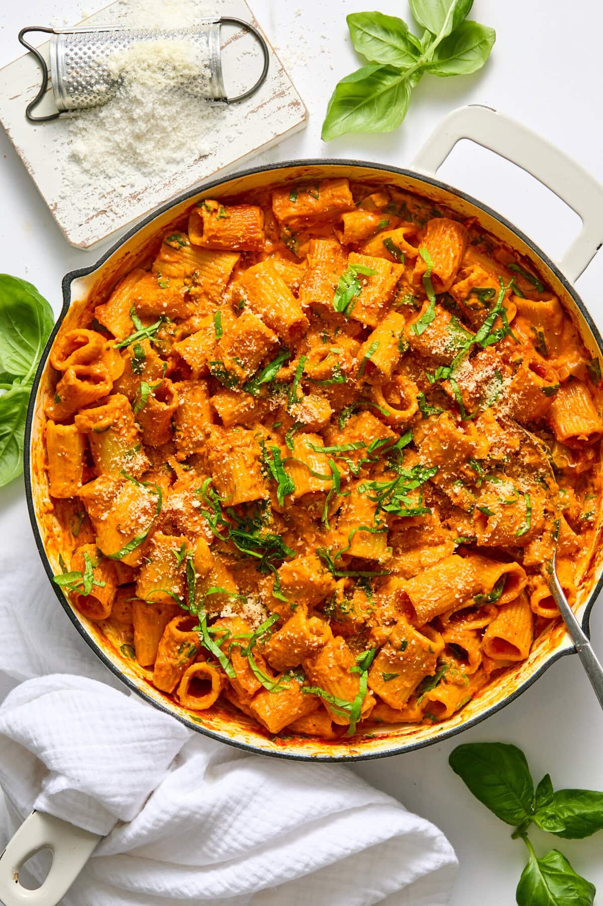 A skillet filled with creamy tomato rigatoni pasta, topped with grated cheese and fresh basil, makes for an irresistible vegan creamy pasta bake. A cheese grater, parmesan, basil leaves, and a white towel sit nearby on a white surface&mdash;perfect props to show how to cook pasta bake in oven for an easy vegan dinner.
