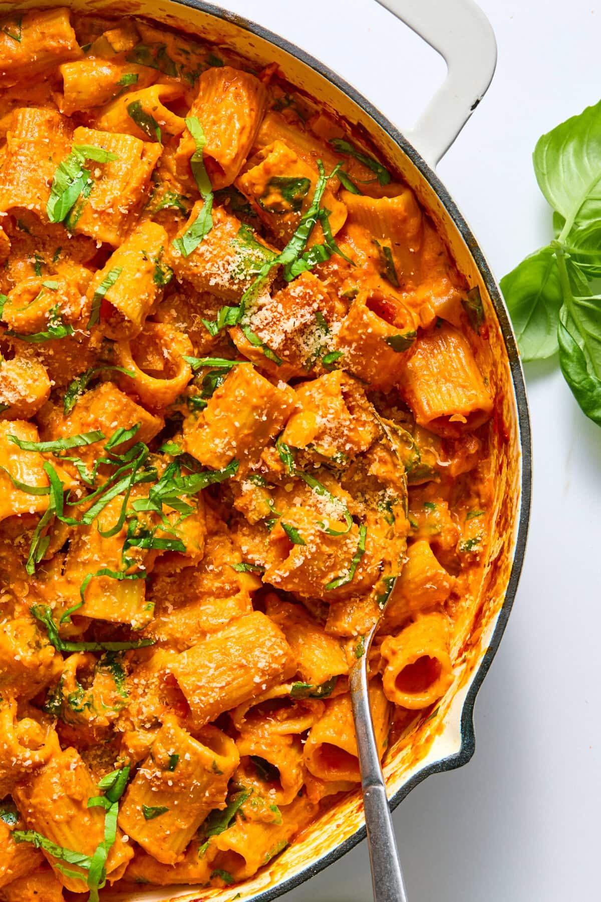 A skillet filled with creamy rigatoni pasta in a tomato sauce, garnished with chopped basil and grated cheese. Perfect for those curious about how to cook pasta bake in oven, this dish features a spoon resting in the pasta and fresh basil leaves on the side.
