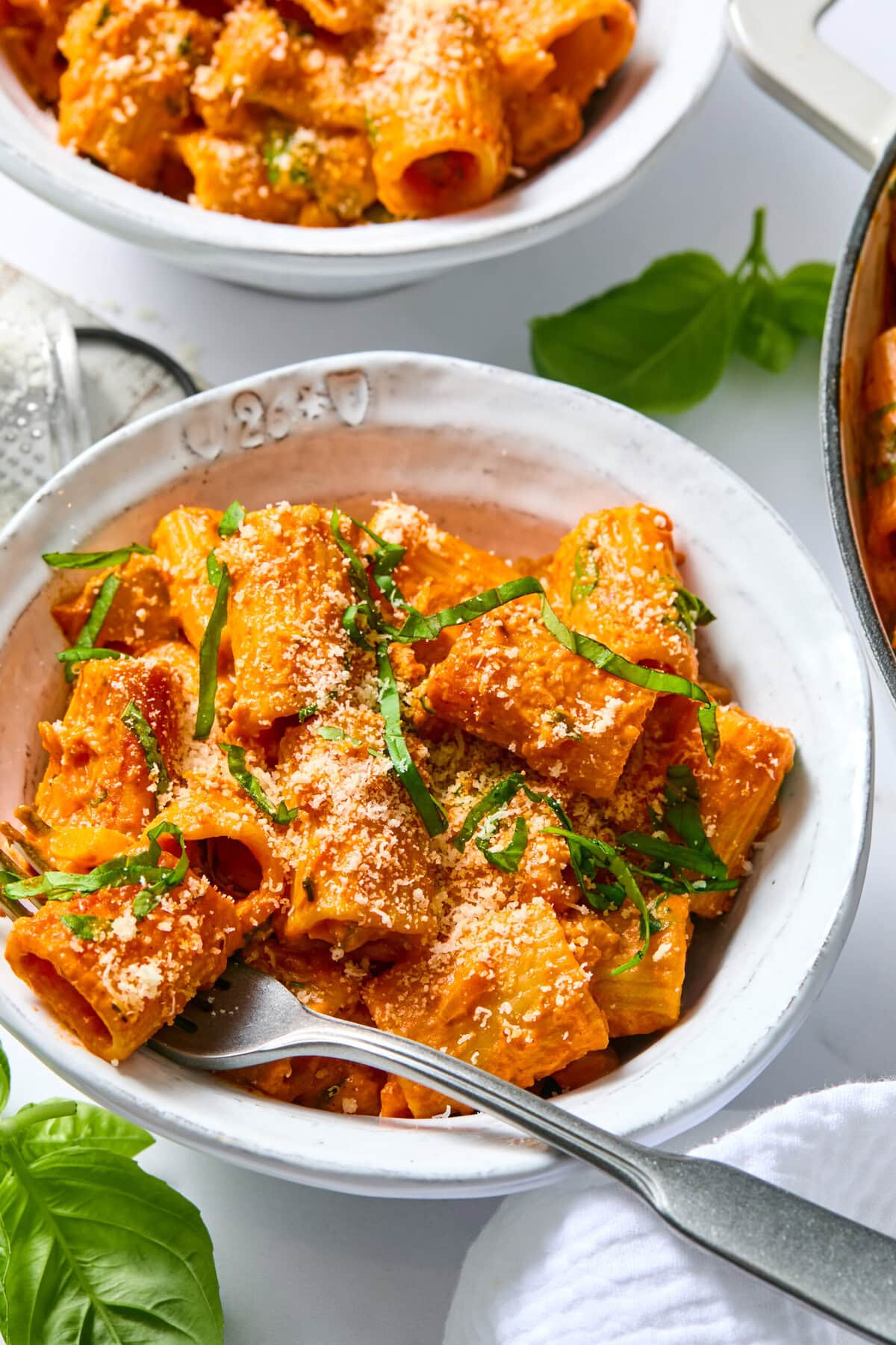 A bowl of rigatoni pasta with creamy tomato sauce, grated cheese, and fresh basil. This delicious vegan creamy pasta bake is served with a fork resting in the bowl, while more pasta and basil leaves are visible in the background.