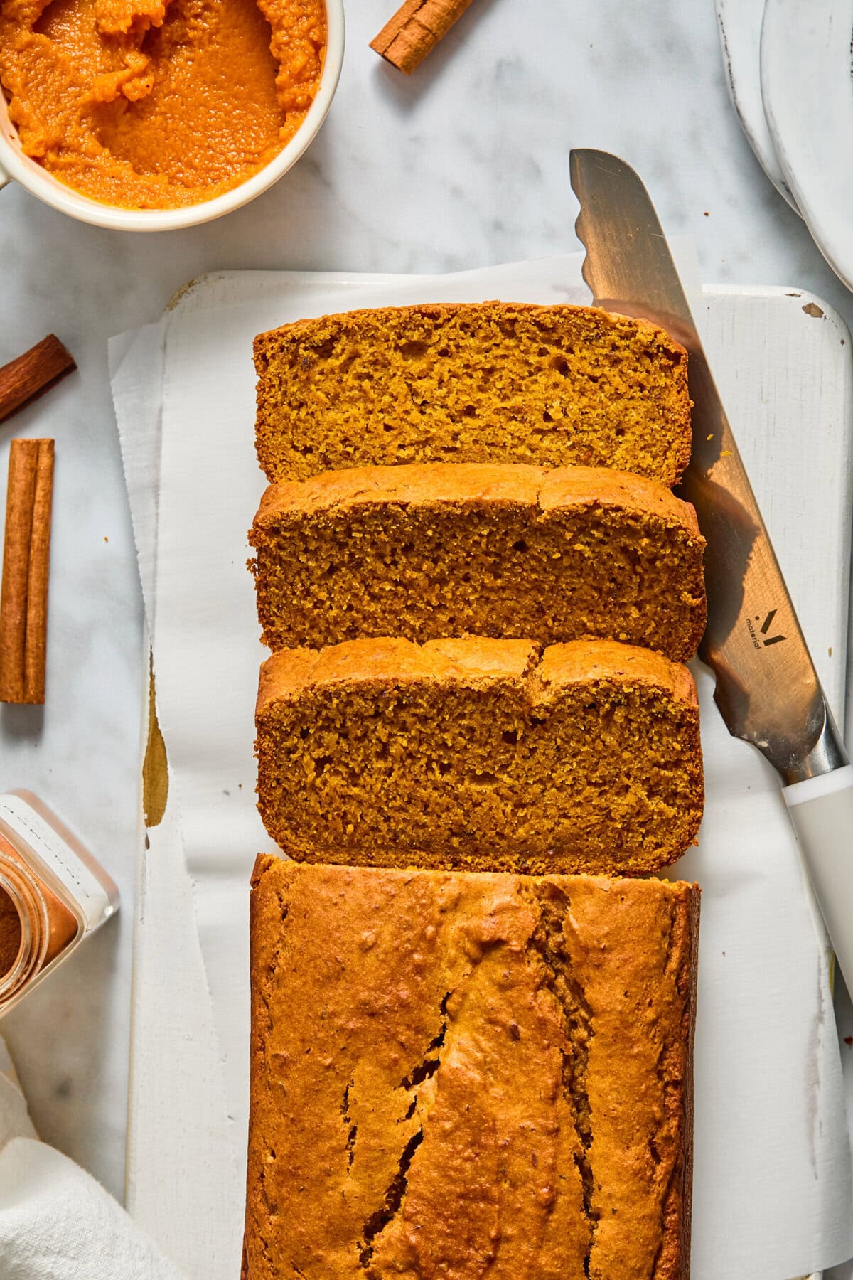 Sliced Vegan Pumpkin Bread on a white cutting board with a knife, a bowl of pumpkin puree, cinnamon sticks, and a spice jar nearby on a marble surface.