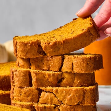 A hand lifts a slice of vegan pumpkin bread from a stack of thick, golden-brown slices on a white surface, with cinnamon sticks nearby and a blurred background.