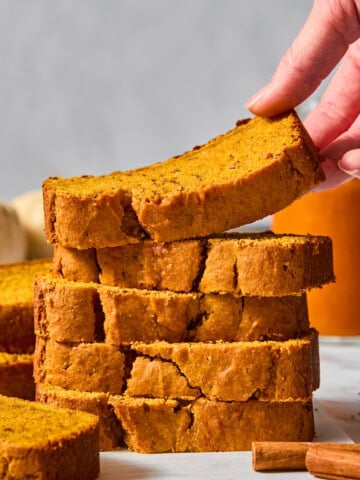 A hand lifts a slice of vegan pumpkin bread from a stack of thick, golden-brown slices on a white surface, with cinnamon sticks nearby and a blurred background.