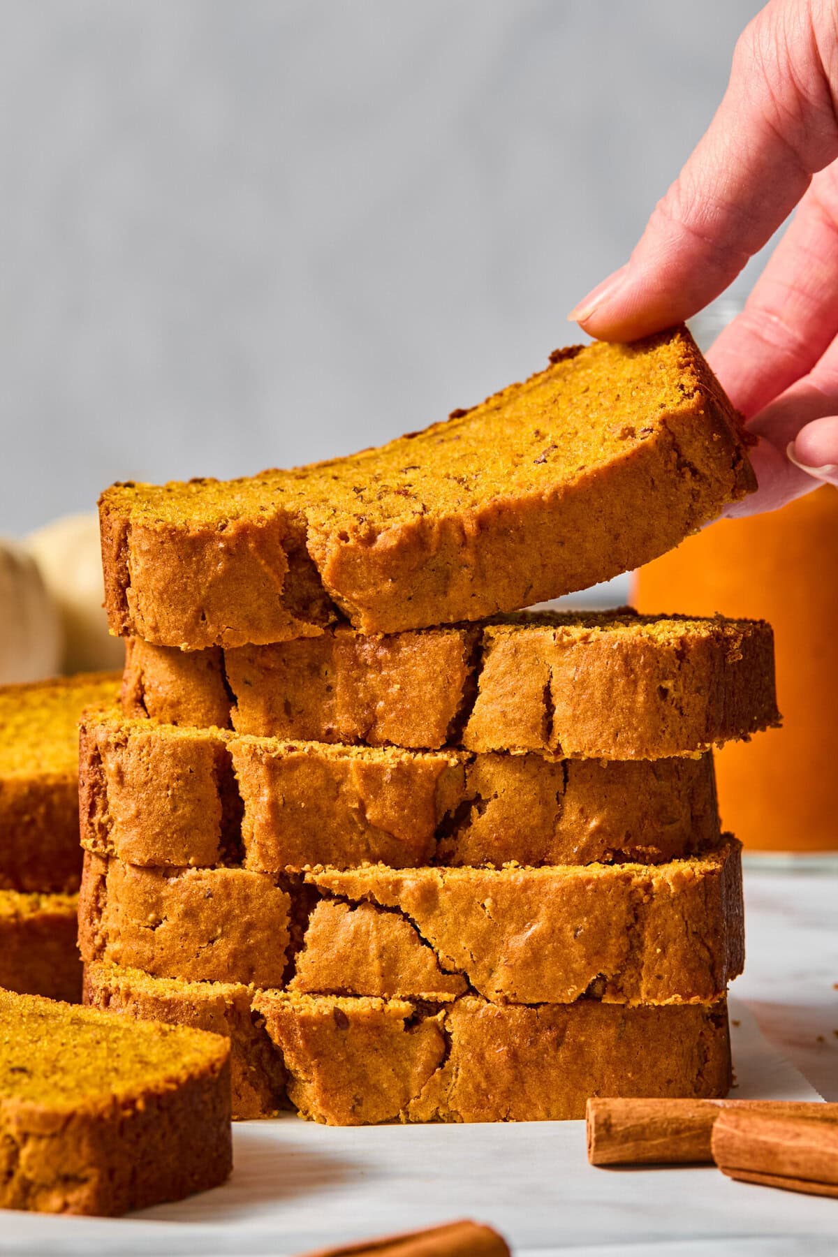 A hand lifts a slice of vegan pumpkin bread from a stack of thick, golden-brown slices on a white surface, with cinnamon sticks nearby and a blurred background.