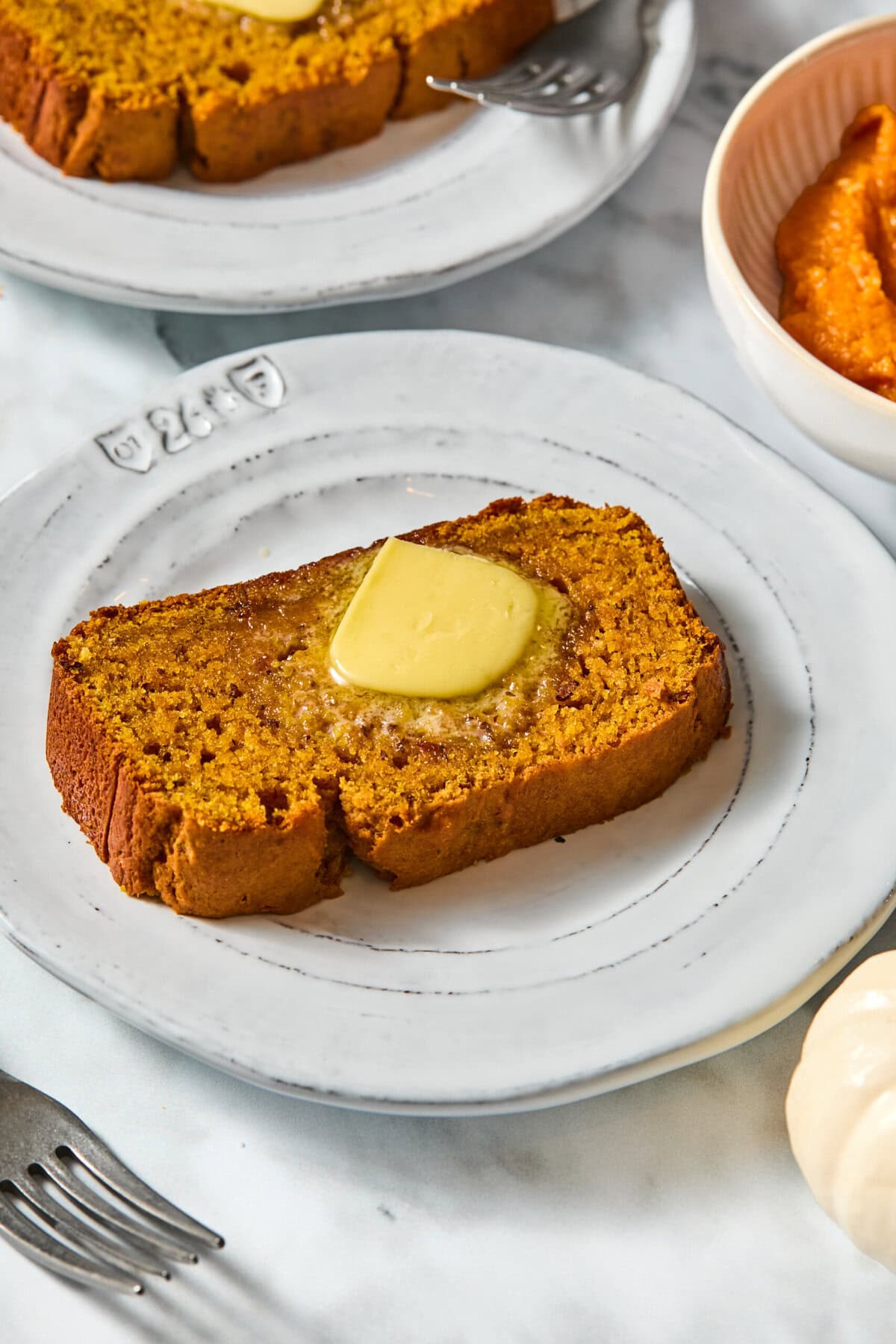 A slice of Vegan Pumpkin Bread with a pat of melting butter on a white plate, accompanied by a fork. Nearby is a bowl of orange puree.